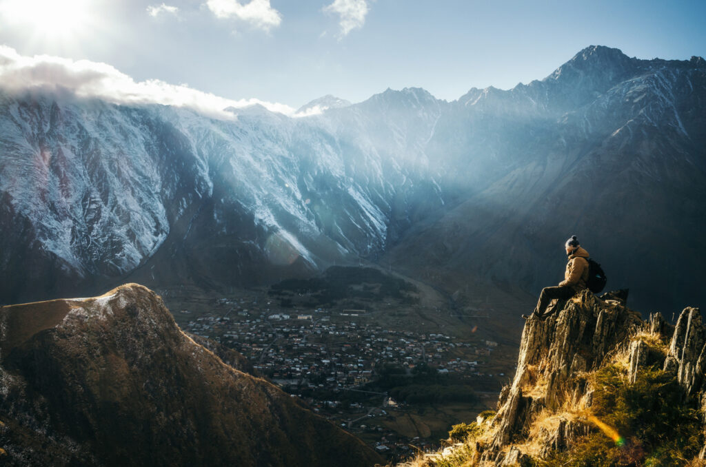 Young tourist in bright hat, black trousers with a backpack sit on cliff's edge and looking at the misty mountain village and glacier at sunrise, Stepantsminda, Georgia